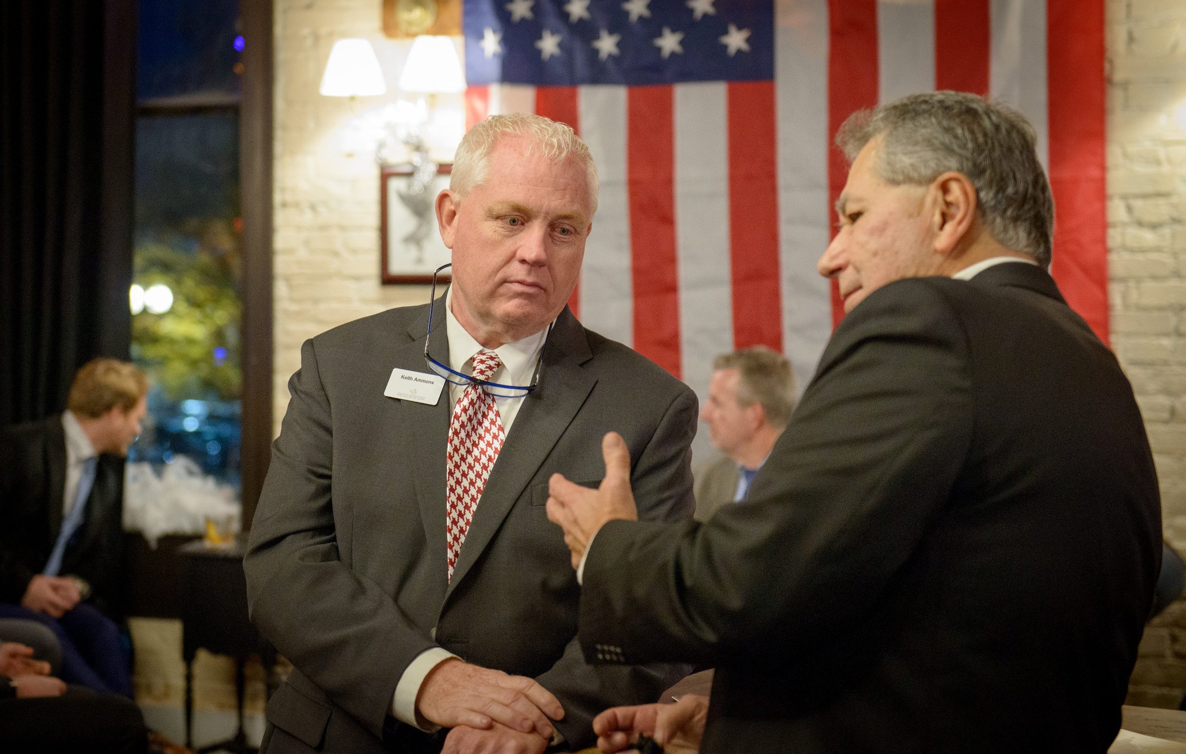 Two Christian business leaders in serious conversation at the CEA Policy Summit, with a large American flag in the background.